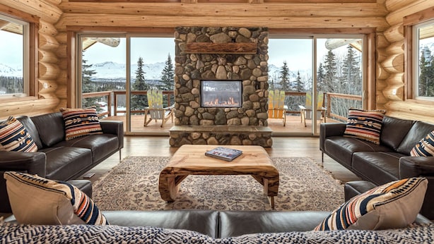 A log cabin interior with a stone fireplace, leather sofas, and a view of snow-capped mountains. - Majestic Valley Wilderness Lodge (Sutton)
