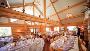 A spacious log cabin dining area with multiple tables set for guests, wooden beams on the ceiling, and large windows allowing natural light. - Majestic Valley Wilderness Lodge (Sutton)