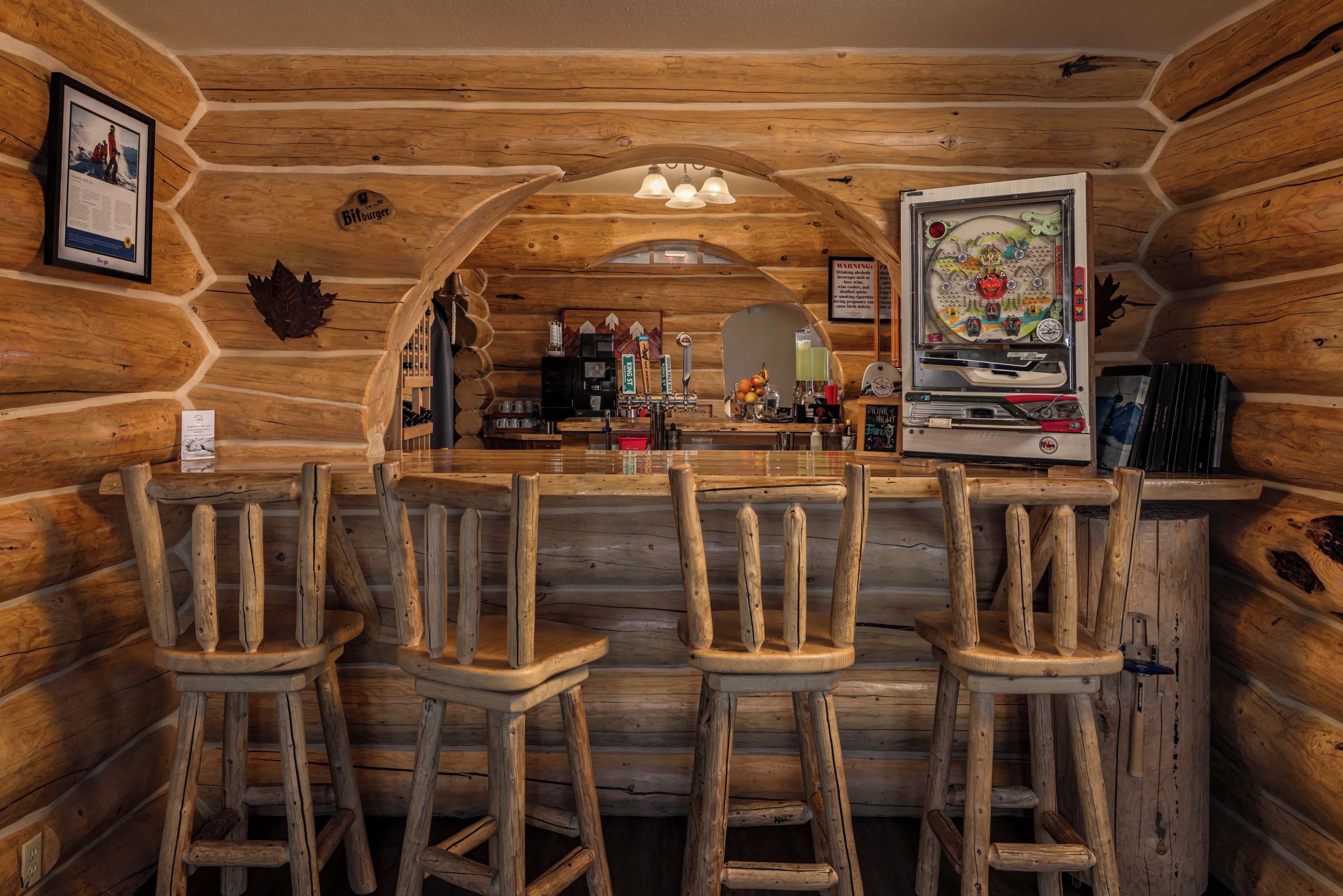 Interior de una cabaña de troncos con una zona de bar que incluye taburetes de madera, una máquina de café y una máquina recreativa de pinball decorativa de gran tamaño.