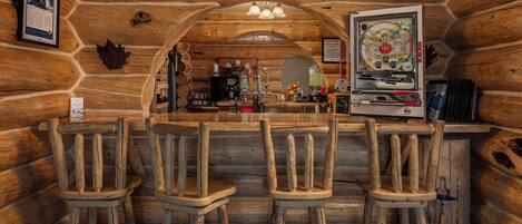 Interior de una cabaña de troncos con una zona de bar que incluye taburetes de madera, una máquina de café y una máquina recreativa de pinball decorativa de gran tamaño.
