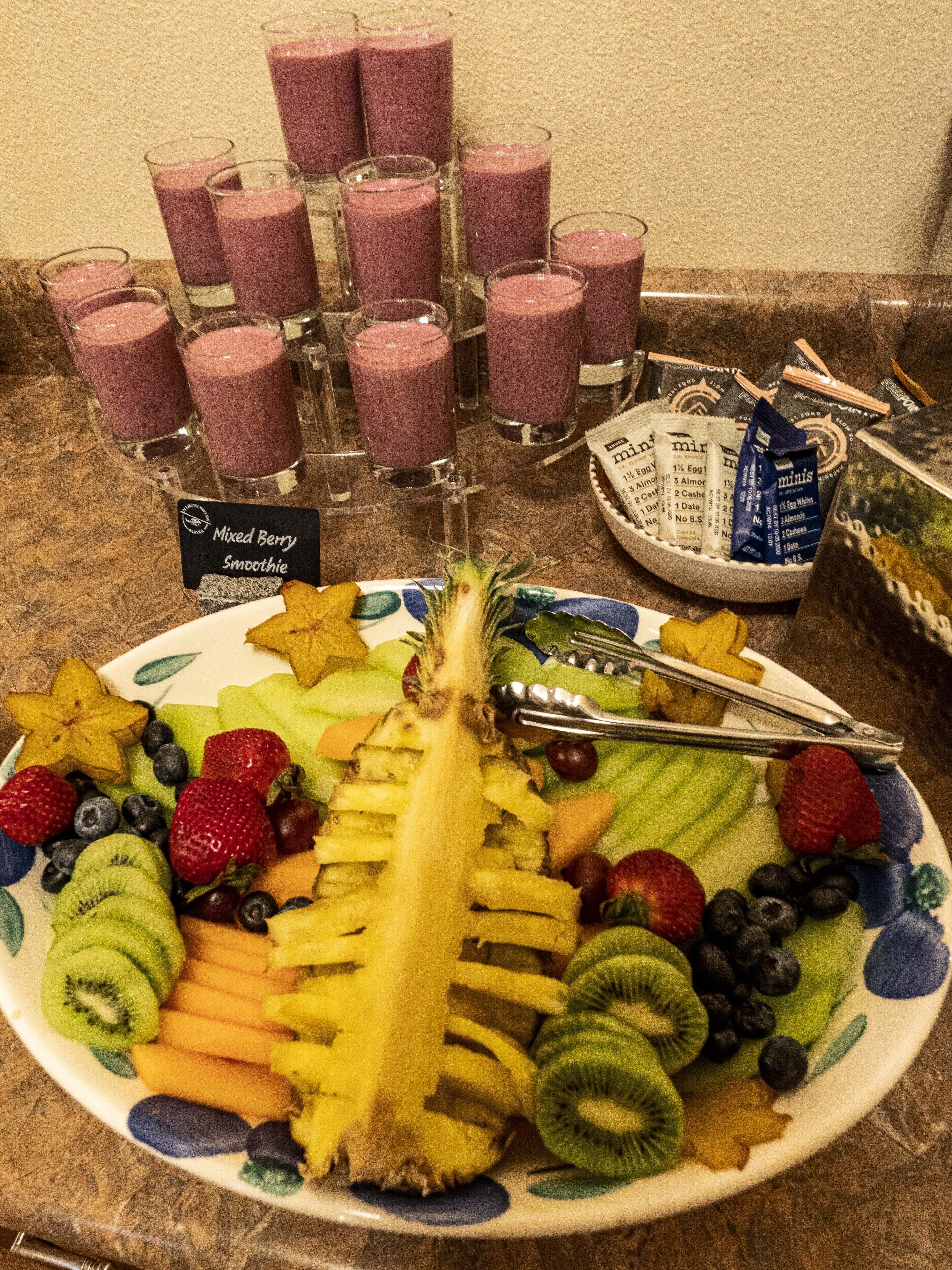 Un plateau de fruits comprenant une variété de fruits coupés en tranches, accompagné d’une pancarte indiquant qu’il y a aussi des smoothies aux fruits rouges.