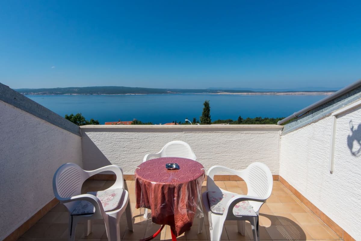 Una terraza con una mesa redonda y cuatro sillas, con vistas al mar tranquilo y colinas lejanas.
