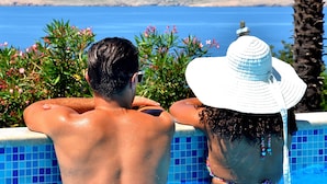 Eine Person lehnt am Poolrand und blickt auf das Meer und eine entfernte Küste.