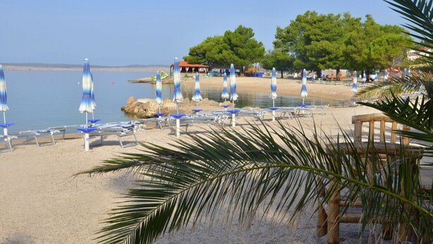 Ein Strand mit Reihen von Sonnenschirmen und Liegestühlen, ein sandiger Uferstreifen und ein Gebäude im Hintergrund.