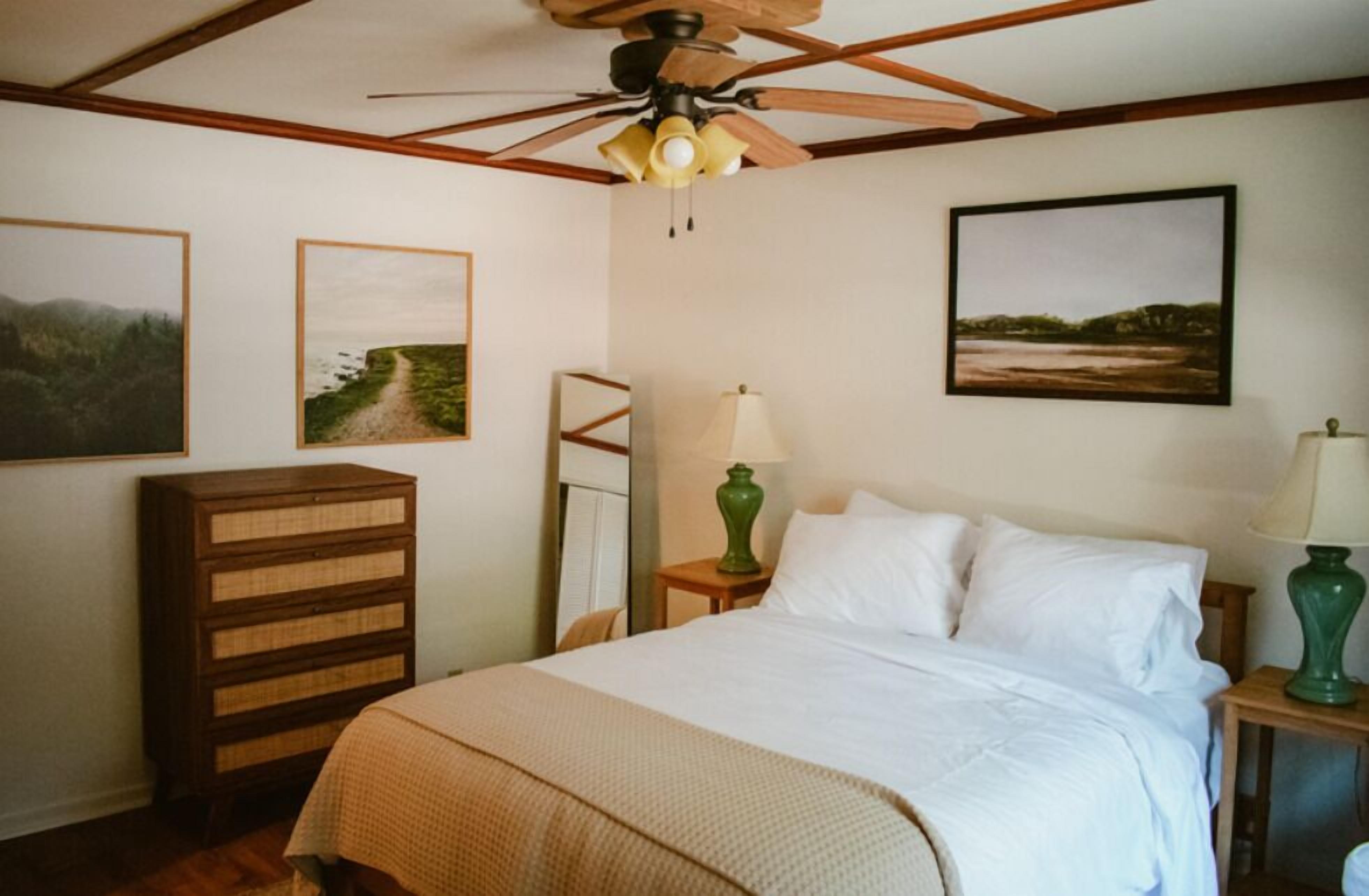 A bedroom with a bed, a wooden dresser, a ceiling fan, and two wall-mounted lamps.