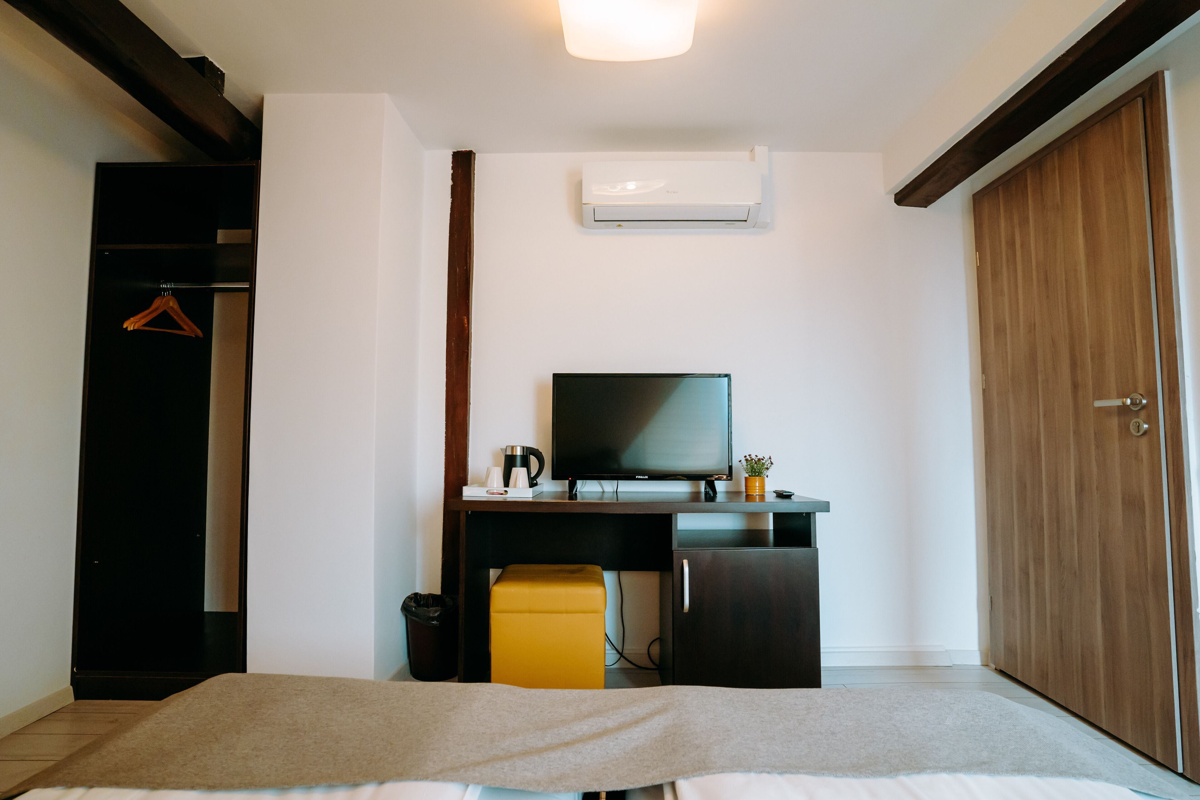 Ein Schlafzimmer mit einem Bett, einem Schreibtisch mit Fernseher, einem kleinen gelben Hocker, einem Kleiderschrank und einer Klimaanlage.