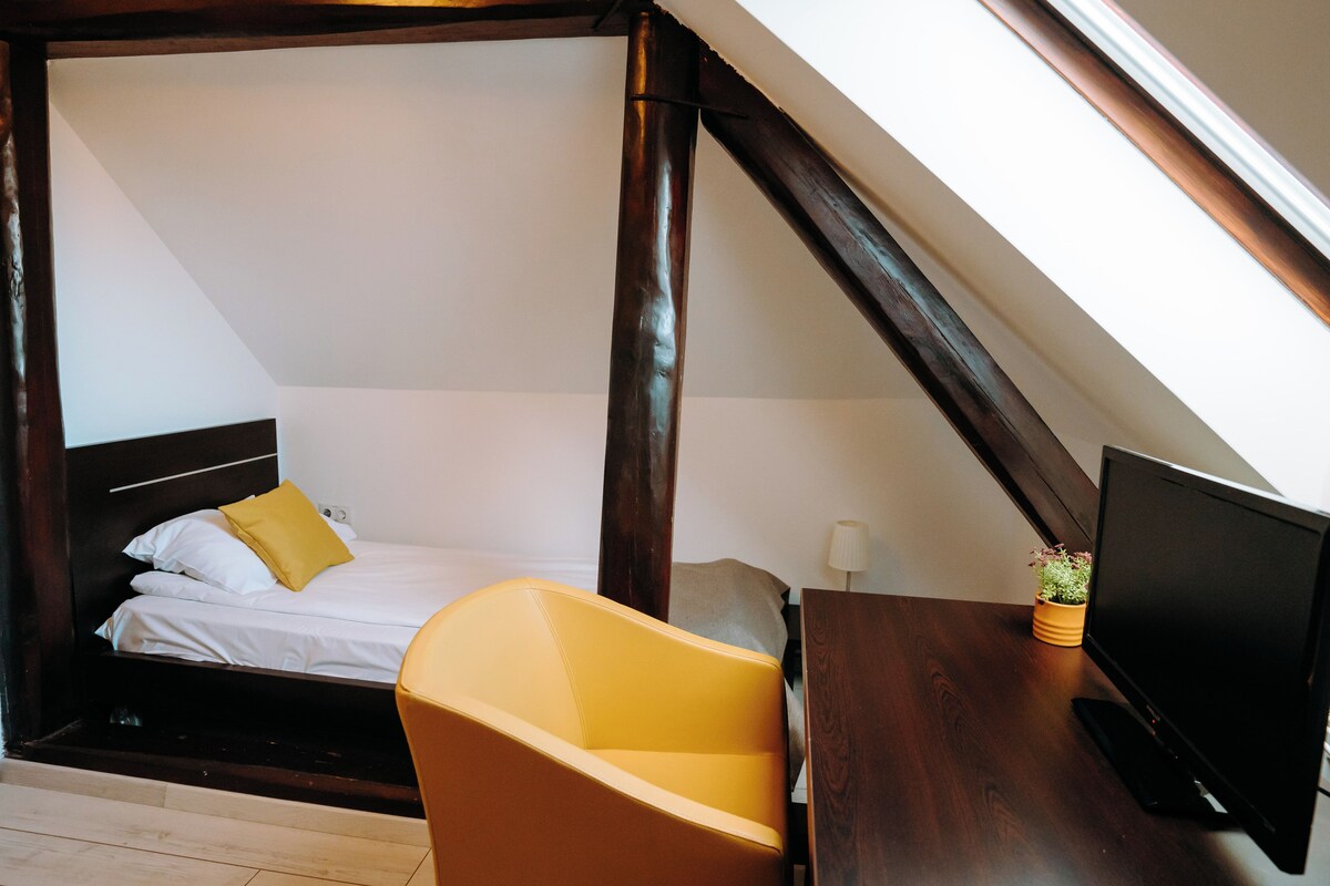 Un dormitorio con una cama, una silla amarilla, un televisor y una planta pequeña sobre una mesa de madera.