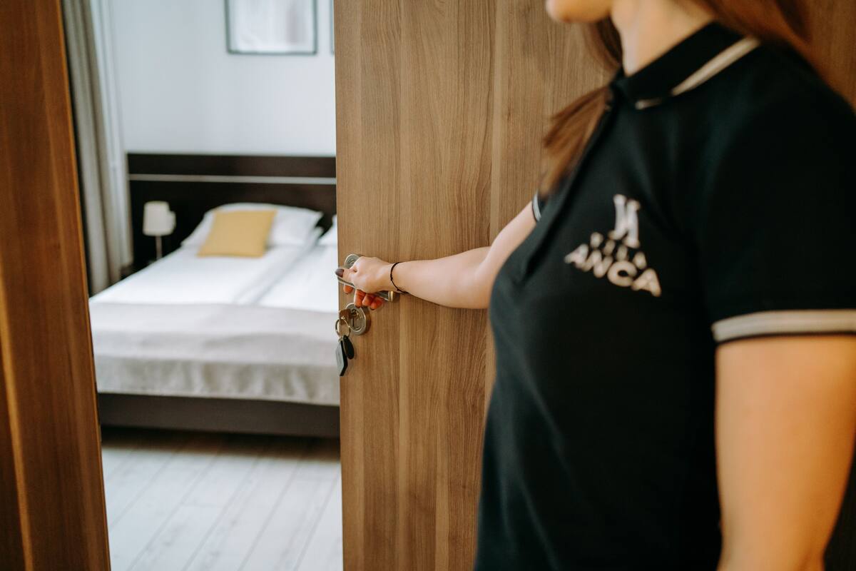 A person holding a key near a wooden door, with a bed and a nightstand visible in the background.