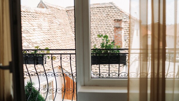 A view through a window with a metal balcony railing, a tiled roof, and some greenery. - Vila Anca (Sibiu)