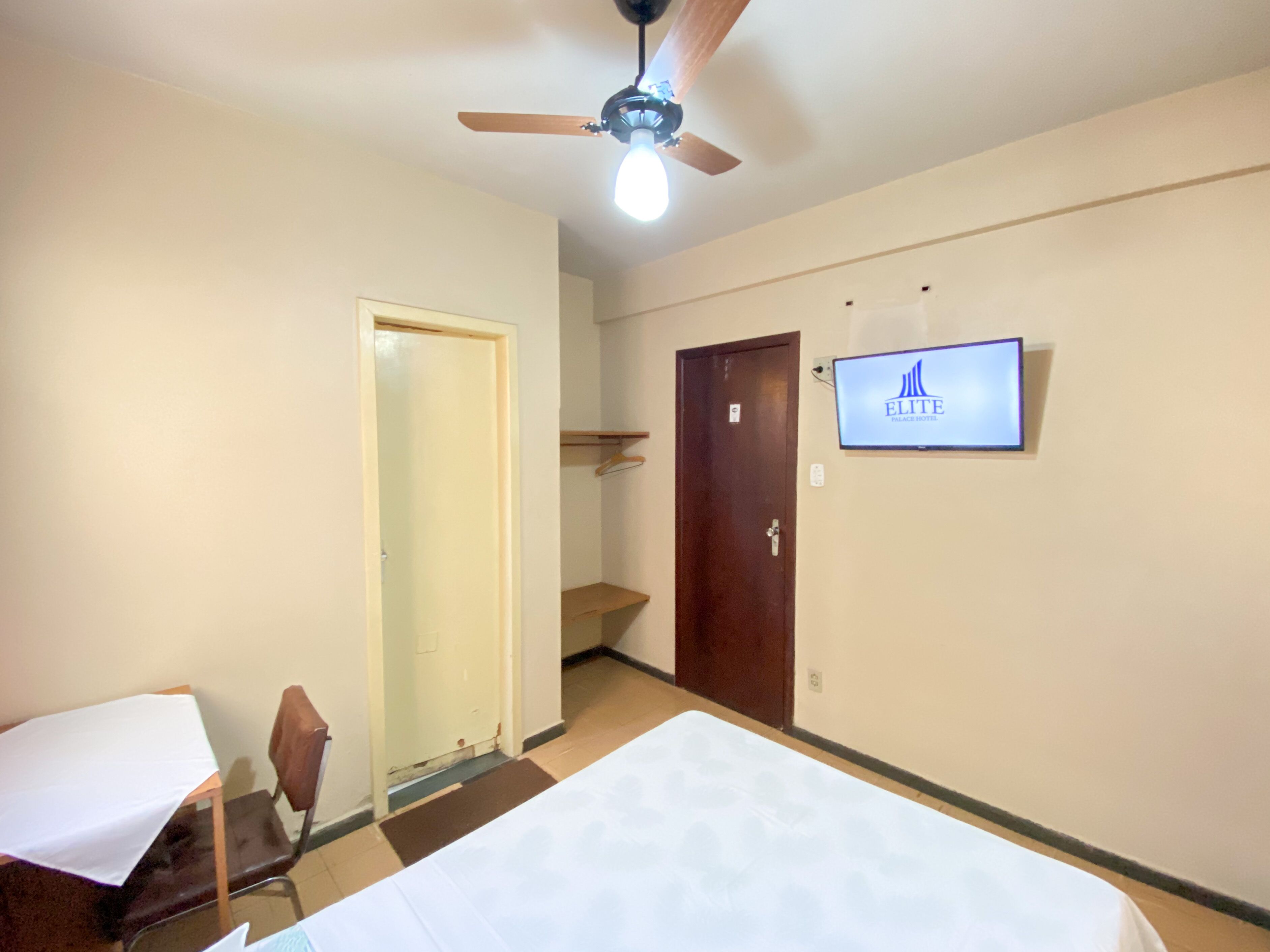 A hotel room with a bed, a desk, a chair, a ceiling fan, a TV, and a door.