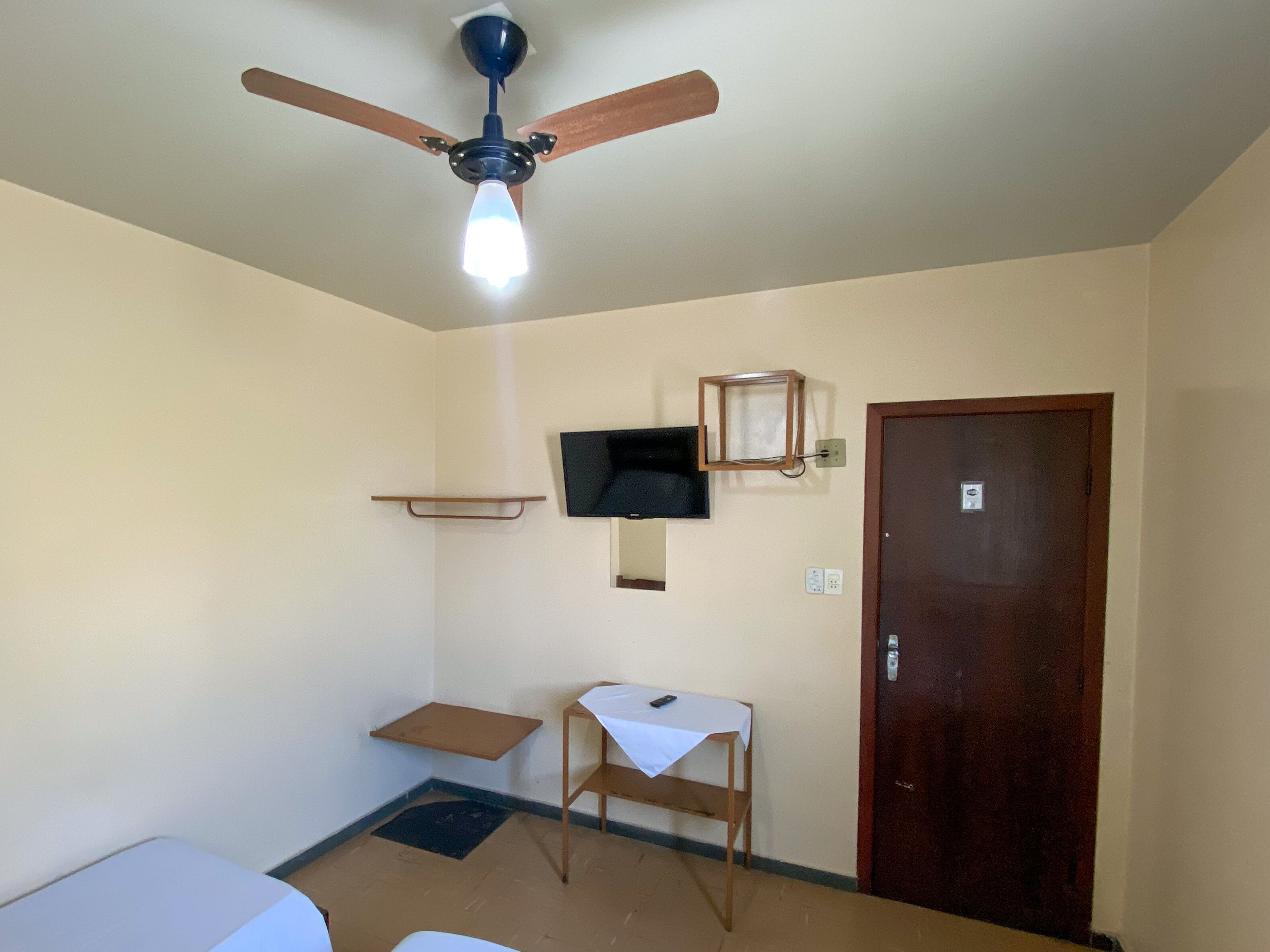 A small, sparsely furnished room with a ceiling fan, a mounted television, a wooden door, and a small table with a white tablecloth.