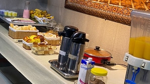 A buffet table with various food items, a coffee maker, a juice dispenser, and a painting on the wall.