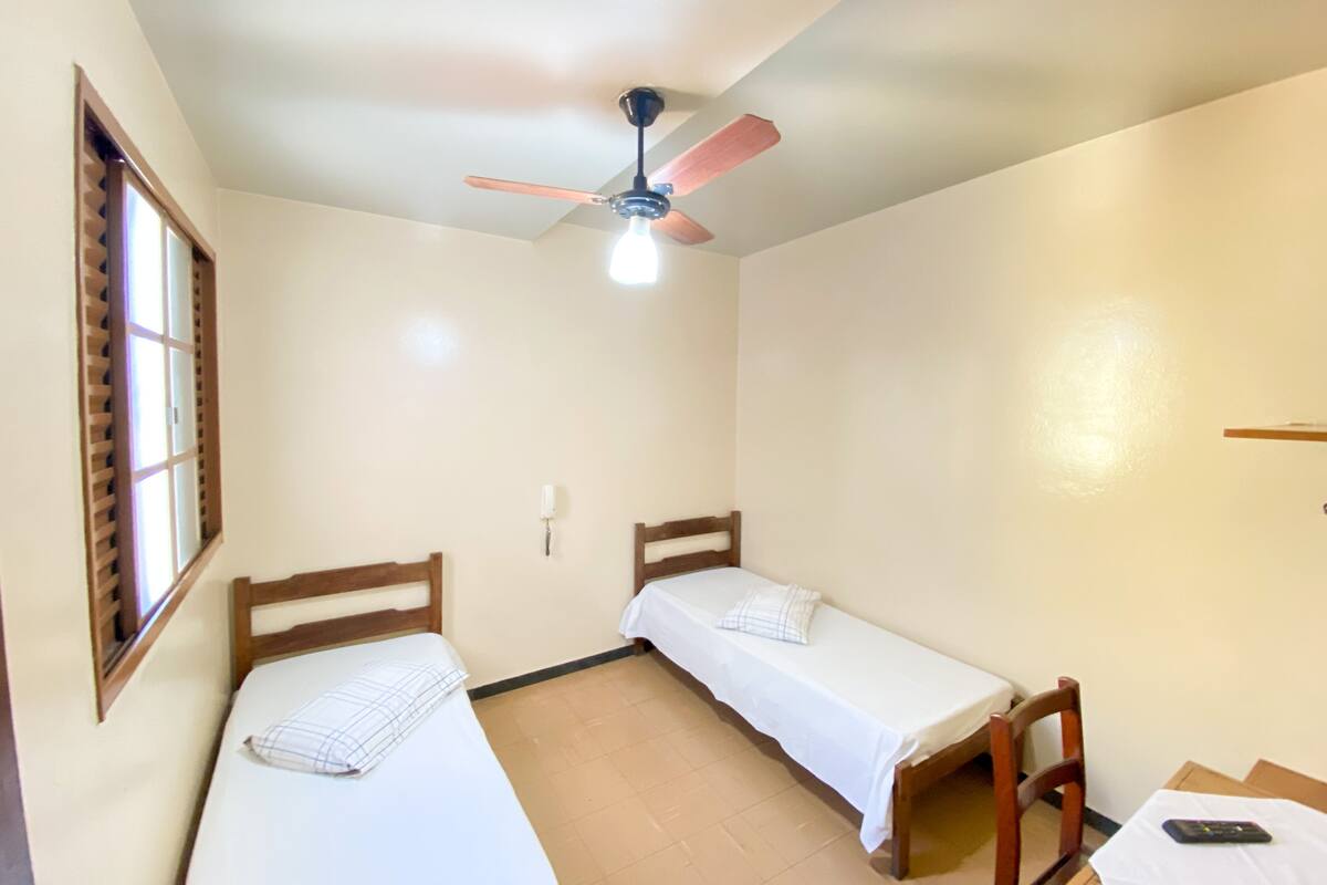 A small, clean room with two single beds, a ceiling fan, a window with blinds, and a small table with a remote control.