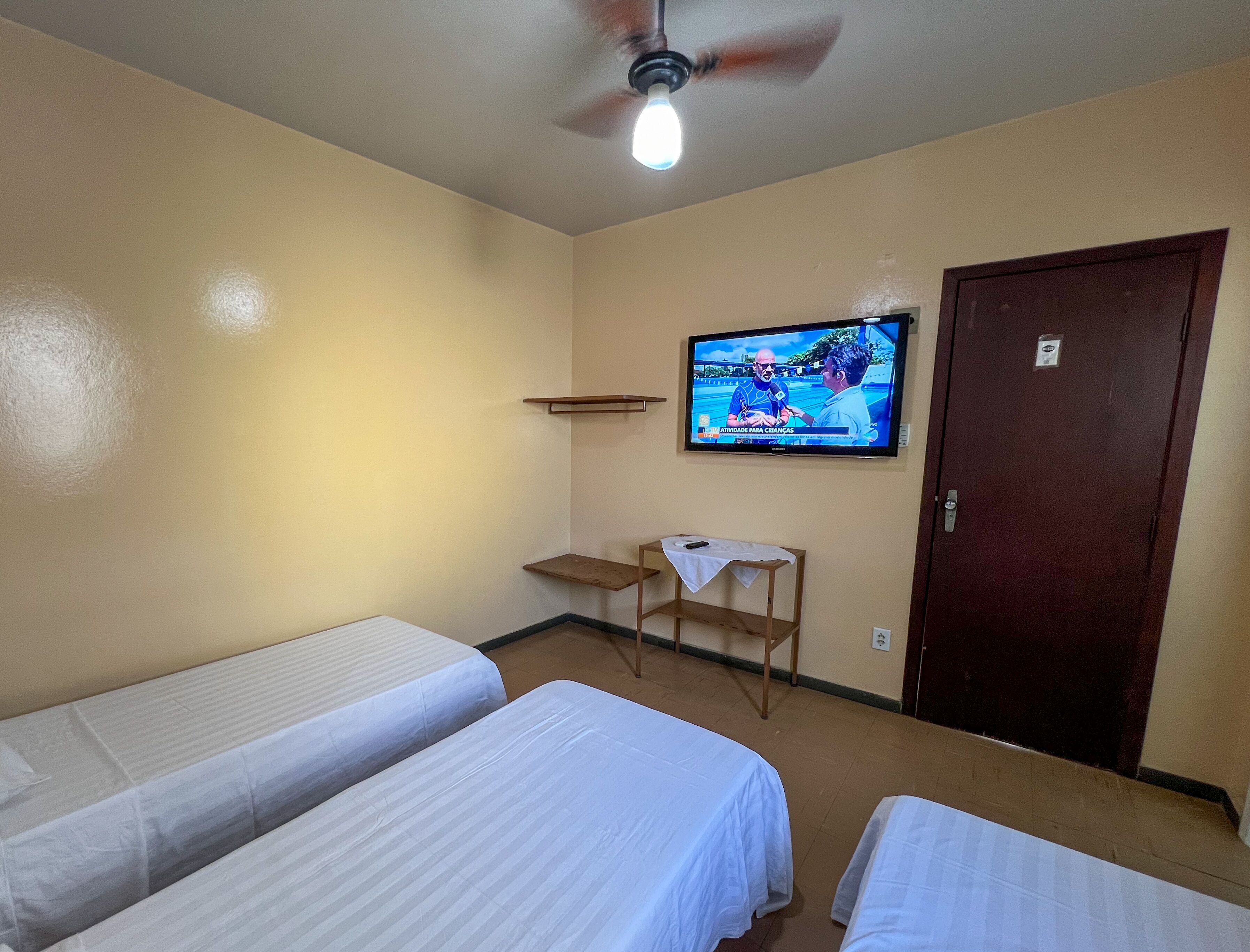 Ein Hotelzimmer mit zwei Einzelbetten, einem an der Wand befestigten Fernseher, einem Deckenventilator und einer Holztür.