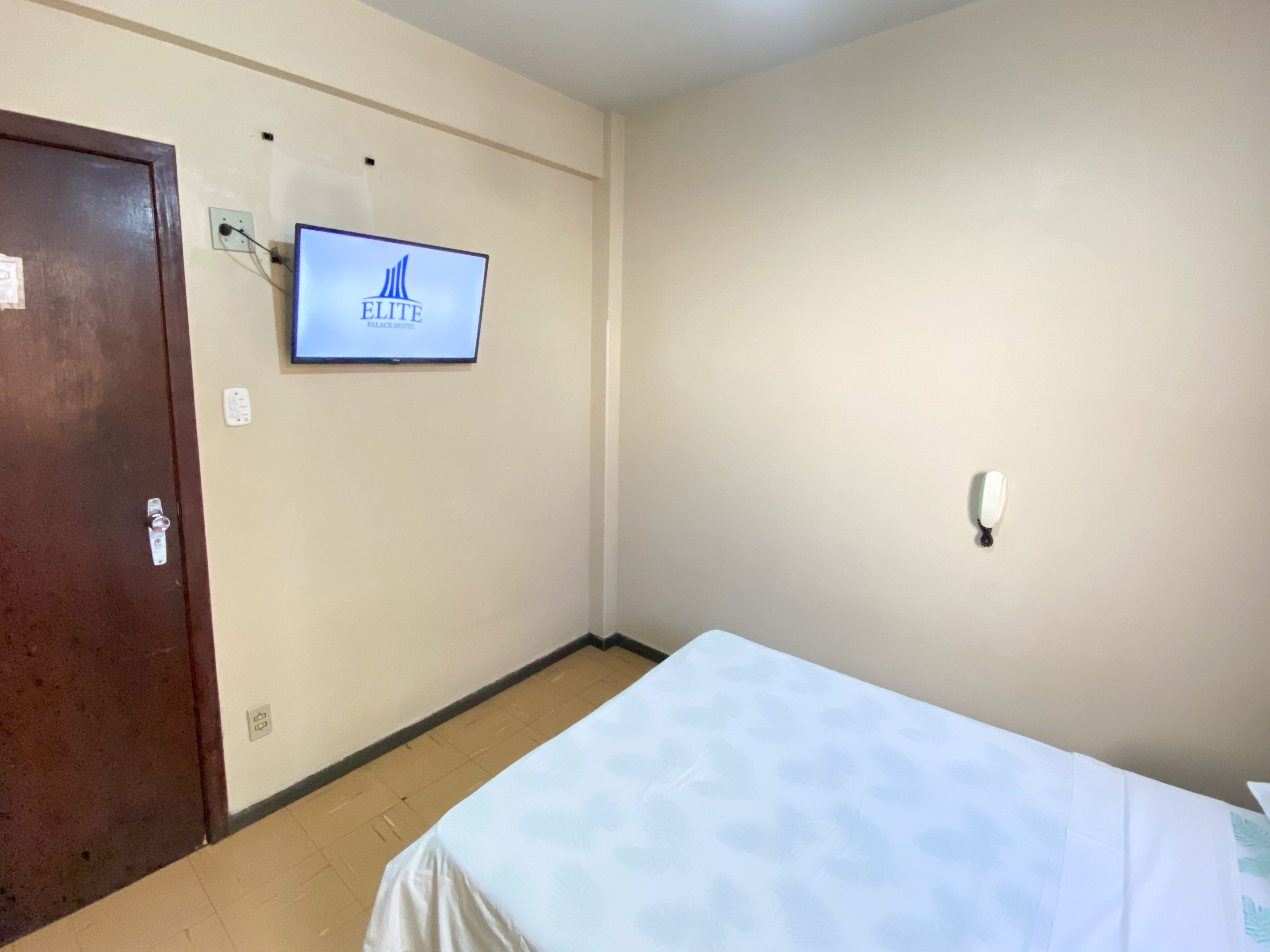Quarto de hotel com uma cama, uma porta, uma televisão fixada na parede exibindo o nome do hotel e um interruptor de luz.