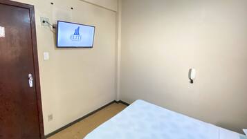 Quarto de hotel com uma cama, uma porta, uma televisão fixada na parede exibindo o nome do hotel e um interruptor de luz.