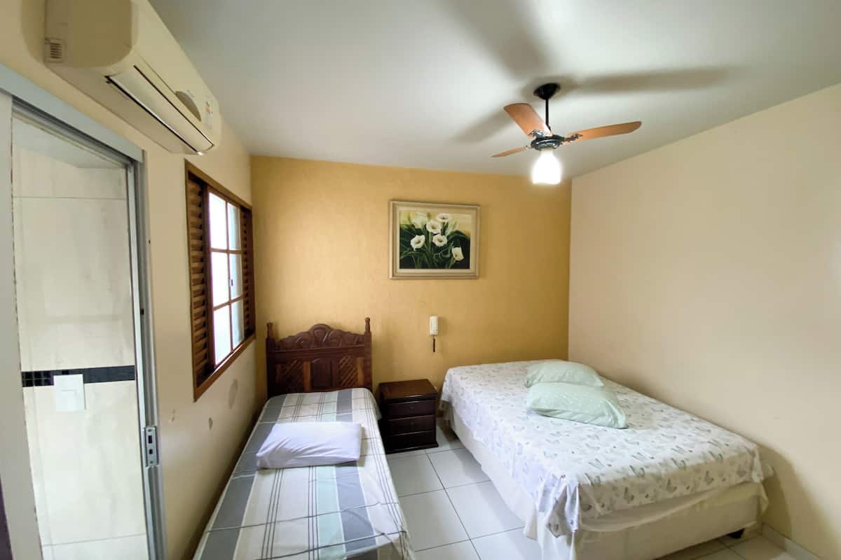 A bedroom with two beds, a ceiling fan, a window with blinds, and a wall-mounted air conditioner.