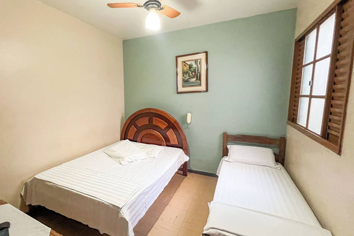 A room with two single beds, a ceiling fan, a framed picture on the wall, and a window with shutters.