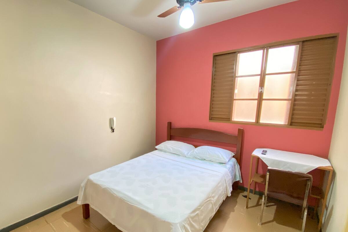 A bedroom with a bed, a desk, a chair, a ceiling fan, and a window with blinds.