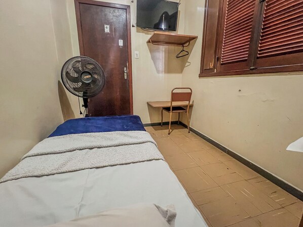 Una habitación pequeña con una cama, un ventilador, una silla, un televisor en una repisa y una ventana con persianas.