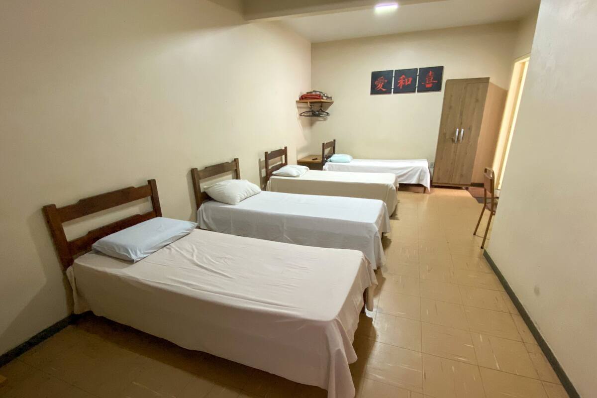 A hotel room with three single beds, a wooden wardrobe, and a wall-mounted shelf with personal items.