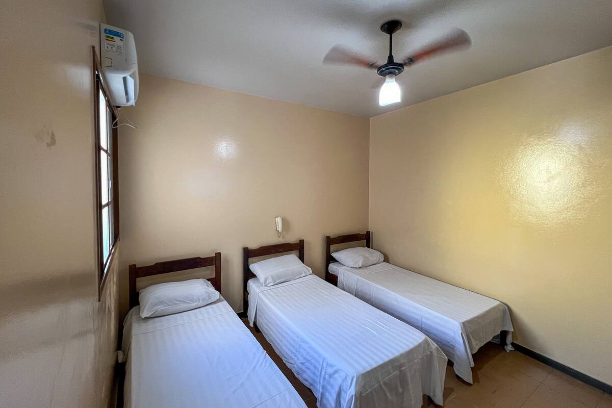 A small, clean room with three single beds, a ceiling fan, and a wall-mounted air conditioning unit.