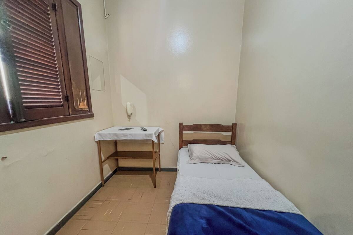A small room with a single bed, a wooden nightstand, a white table, and a window with blinds.