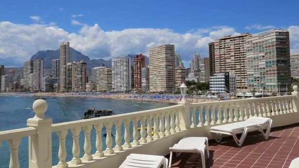A balcony with white lounge chairs overlooking a beach and a cityscape.