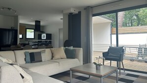 Ein modernes Wohnzimmer mit einer Couch, einem Couchtisch und einem Fernseher. Ein großes Fenster bietet einen Blick ins Freie.