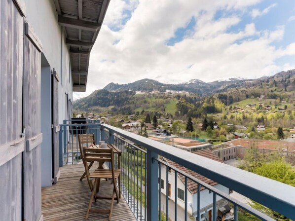A balcony with a wooden table and chairs, overlooking a mountainous landscape with houses and greenery.