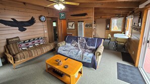 A cozy cabin interior with a wooden ceiling, a ceiling fan, a couch with a blanket, a coffee table, and a dining area with a table and chairs.