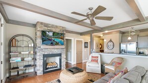 A living room with a stone fireplace, a television mounted above it, and a ceiling fan. - Chalakee Lodge CD-5- Mountain View, Gated Resort, Indoor/Outdoor Pools & Hot Tub (Boone)