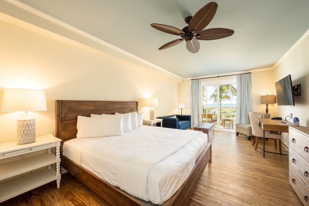 A hotel room with a large bed, wooden headboard, two bedside tables with lamps, a desk with a chair, and a ceiling fan.