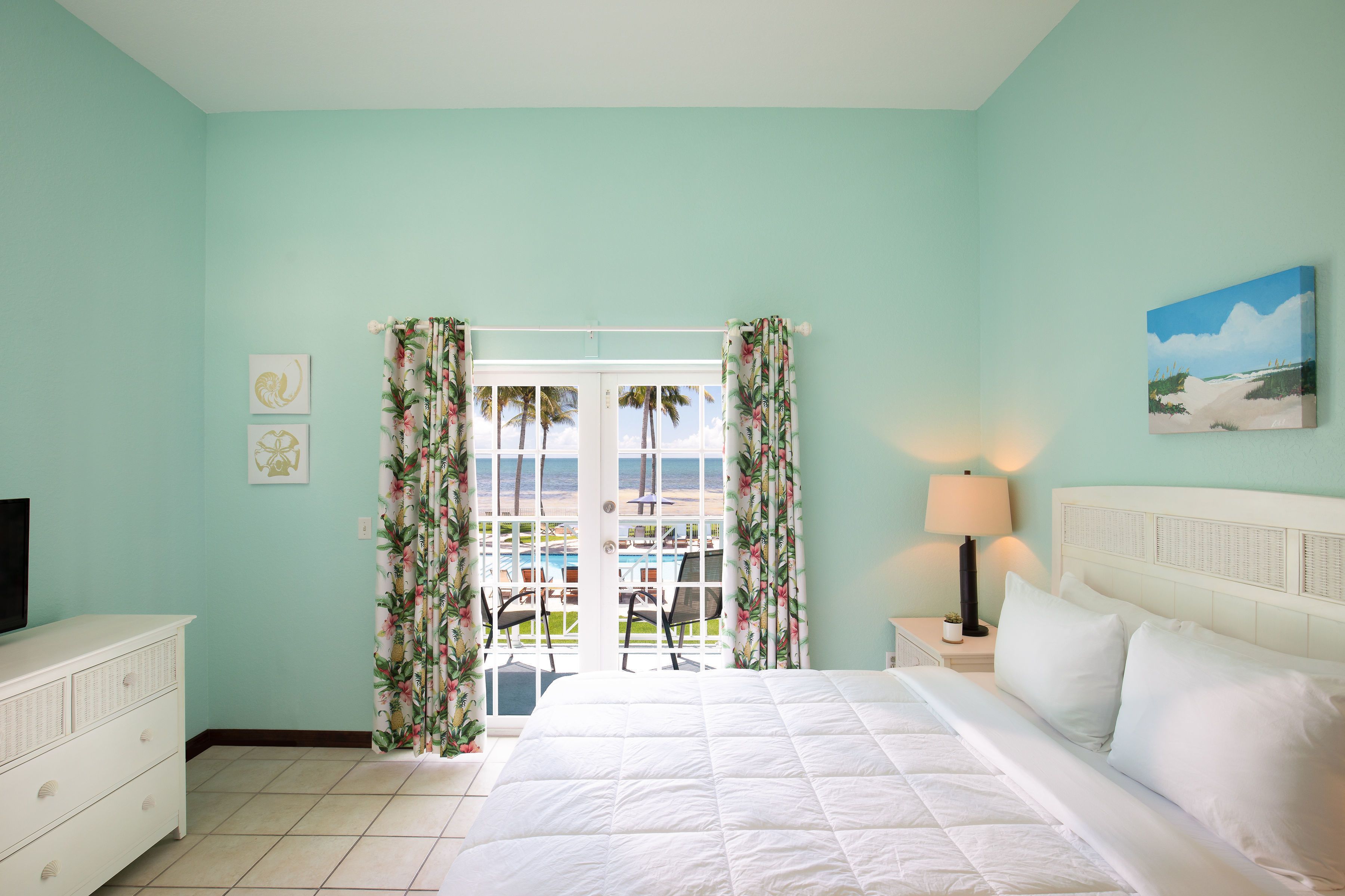 Un dormitorio con cama, televisión, mesita de noche, lámpara y vistas a una piscina y palmeras.