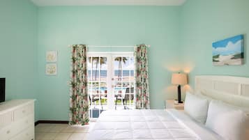 A bedroom with a bed, a TV, a nightstand, a lamp, and a view of a pool and palm trees.