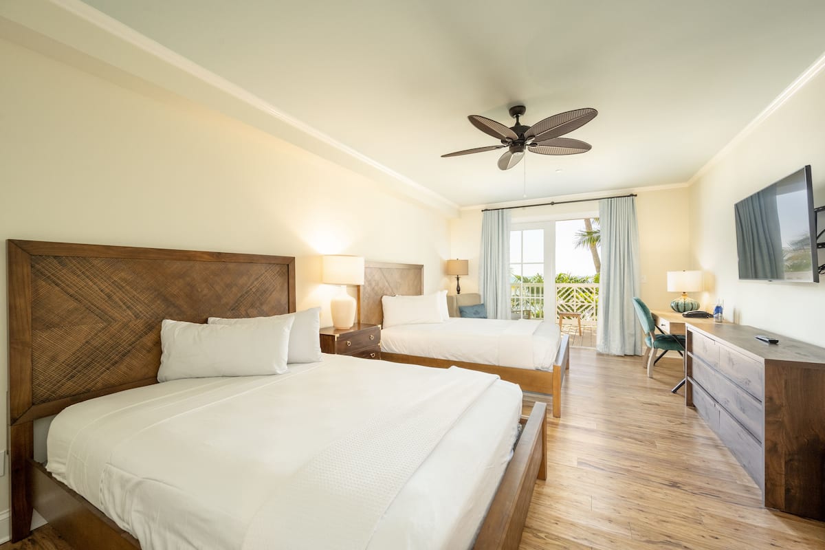 A hotel room with two beds, a desk, a chair, a ceiling fan, and a large window with curtains.