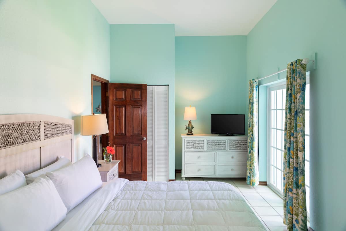 A bedroom with a bed, a nightstand, a lamp, a dresser, a television, and a window with curtains.