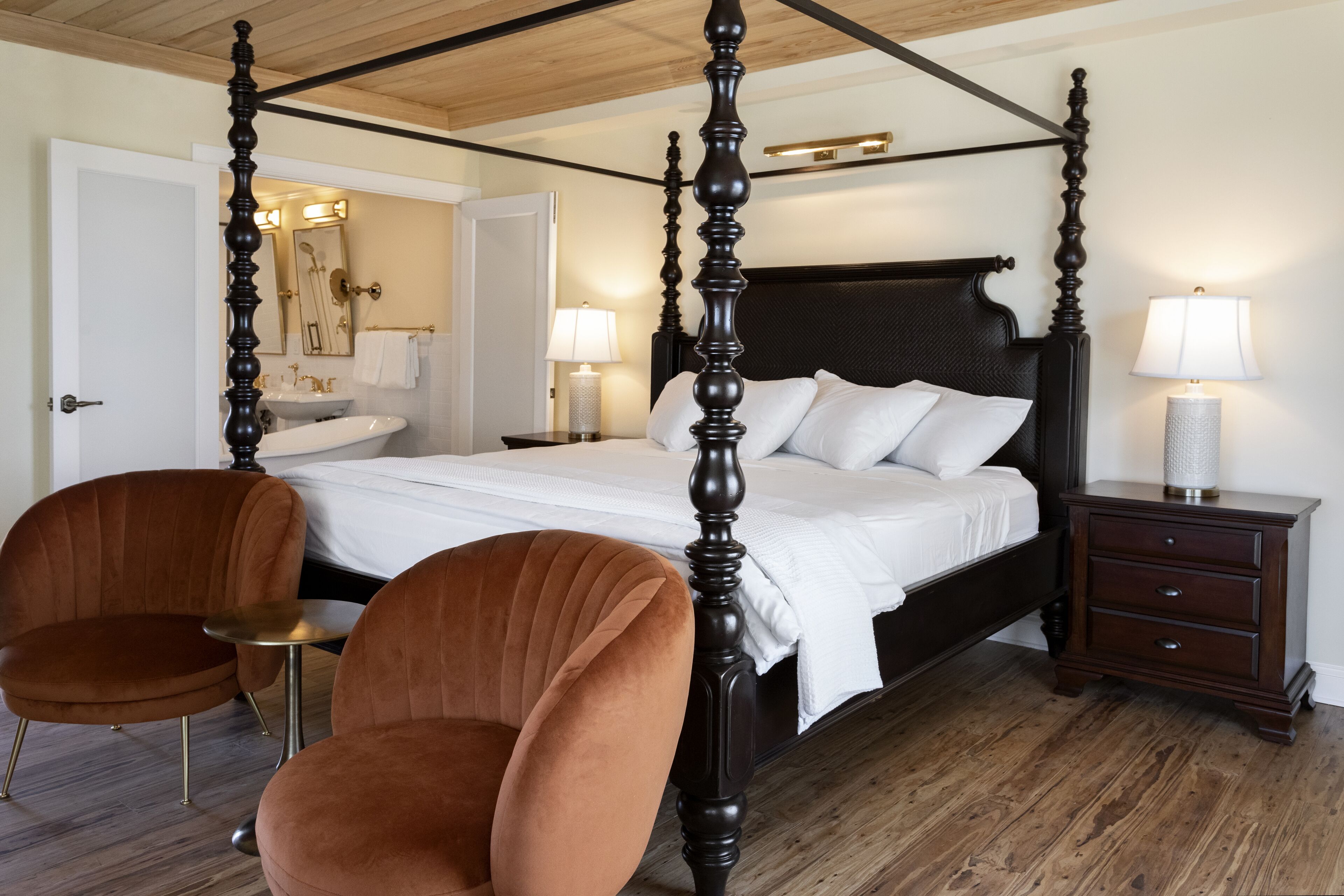 A four-poster bed with a dark wooden frame, a white bedspread, and two white pillows. There is a brown upholstered chair and a small round table beside the bed. A wooden nightstand with a lamp and a drawer is to the right of the bed.
