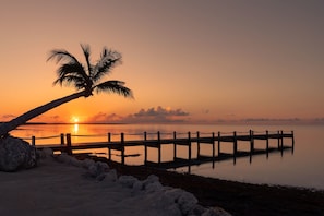 A sunset over a calm body of water with a wooden pier and a palm tree silhouetted against the sky.