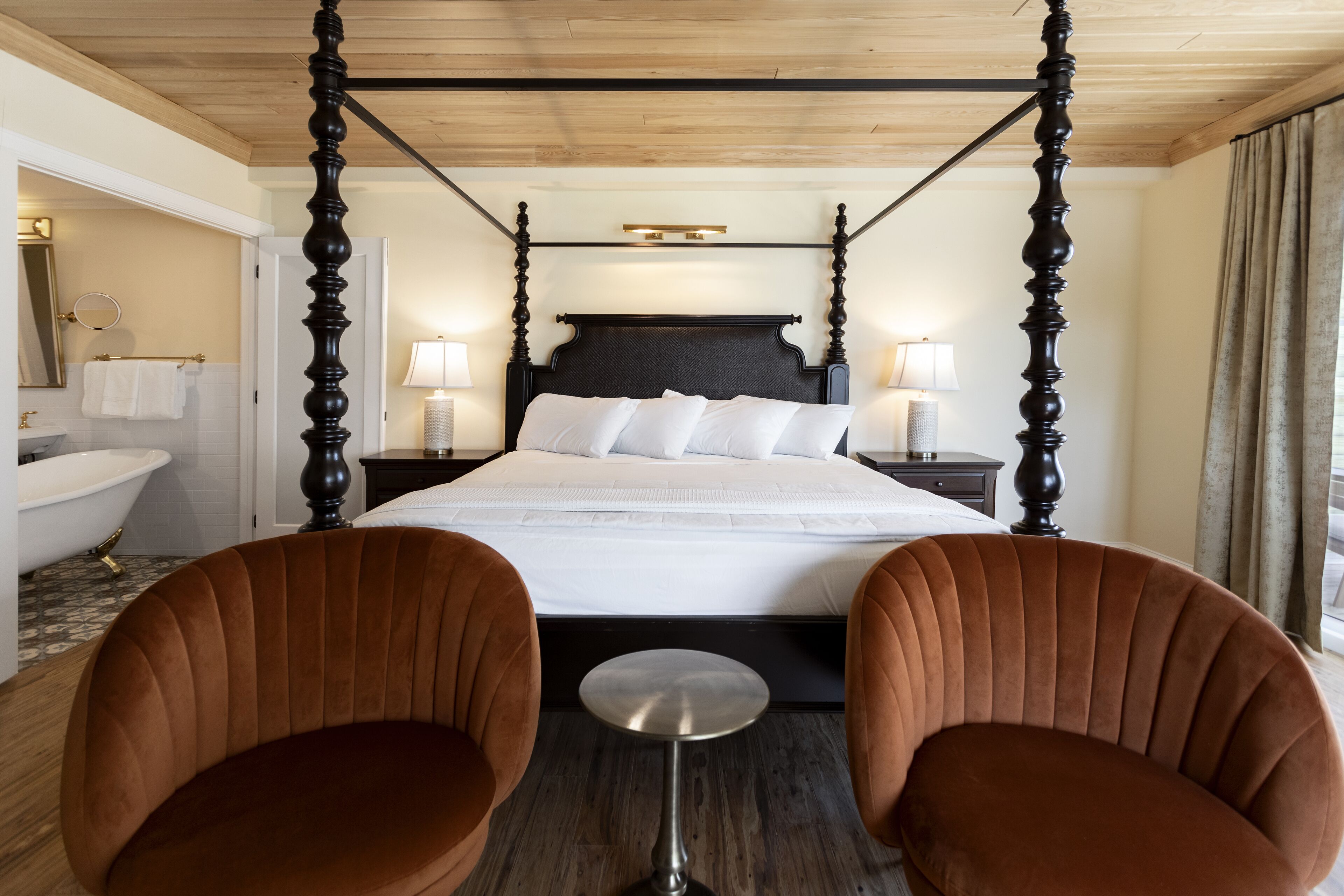 A four-poster bed with a dark headboard, two bedside tables with lamps, a round mirror, a white bathtub, and two brown upholstered chairs.