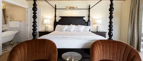 A four-poster bed with a dark headboard, two bedside tables with lamps, a round mirror, a white bathtub, and two brown upholstered chairs.