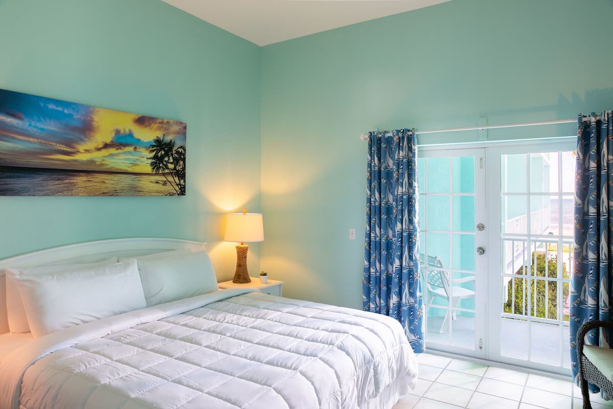 A bedroom with a bed, a nightstand with a lamp, a painting of a sunset, and a view through a door to an outdoor area.