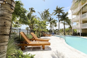 A poolside area with wooden lounge chairs, a swimming pool, palm trees, and a multi-story building.