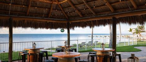 An outdoor dining area with thatched roof, wooden tables, and bar stools, overlooking the ocean.
