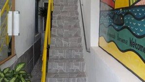 Uma escadaria de pedra com corrimão amarelo que leva a um quarto. Um mural colorido com um sol e ondas está na parede, e uma placa de boas-vindas é visível.