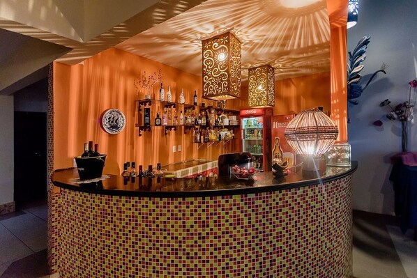 Una zona de bar con mostrador de baldosas, estanterías llenas de botellas y lámparas decorativas colgantes.