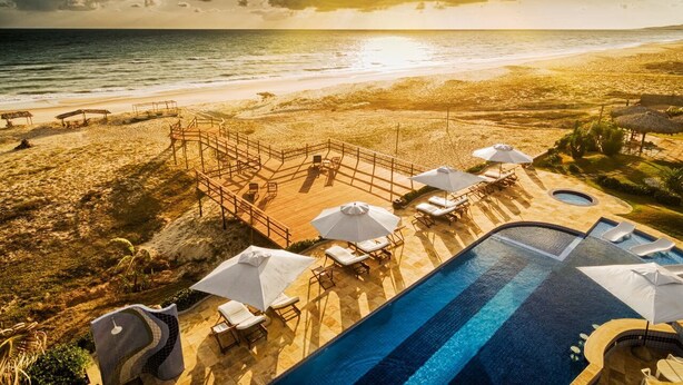 Un espace piscine doté de chaises longues et de parasols, avec vue sur une plage et l’océan.