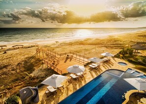 Un espace piscine doté de chaises longues et de parasols, avec vue sur une plage et l’océan.