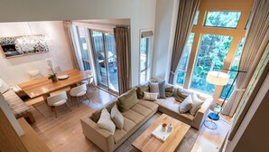 Ein modernes Wohnzimmer mit Couch in Beige, einem Holztisch und einem Essbereich mit Holztisch und weißen Stühlen. Große Fenster sorgen für natürliches Licht und Ausblick ins Freie.