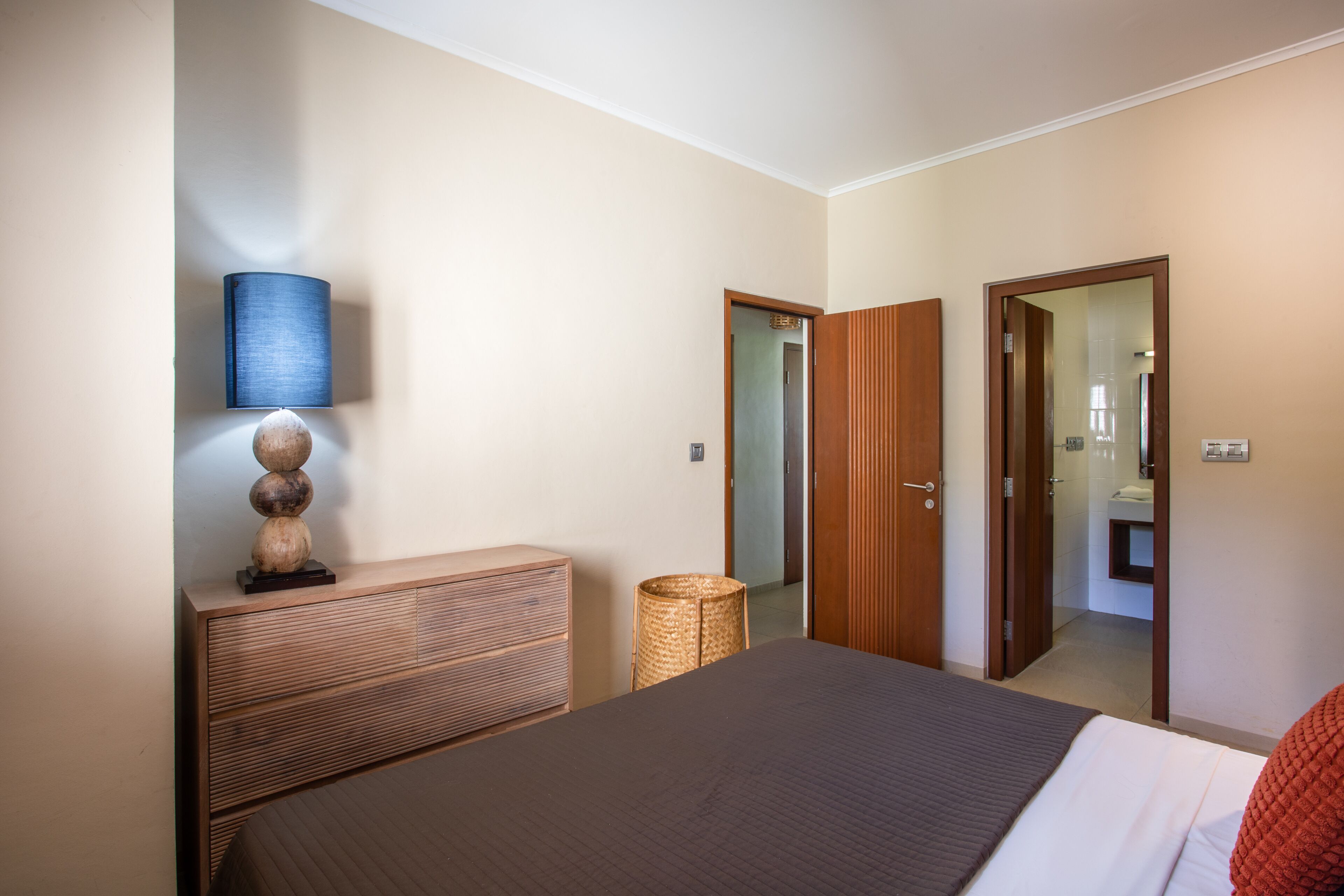 Une chambre à coucher comprenant un lit, une commode, une lampe et une porte donnant sur une salle de bain.